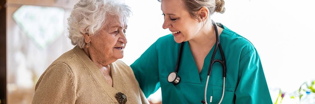 A smiling health professional assists a senior woman with dementia during a care visit.