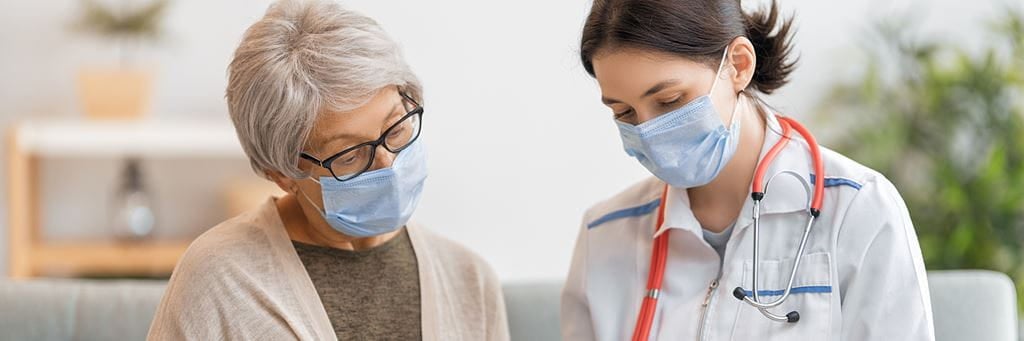 A senior woman in a face mask speaks to a doctor also wearing a face mask.