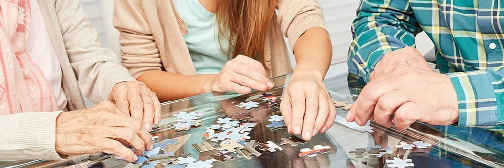 An elderly man and woman and a younger woman do a jigsaw puzzle on a glass tabletop.