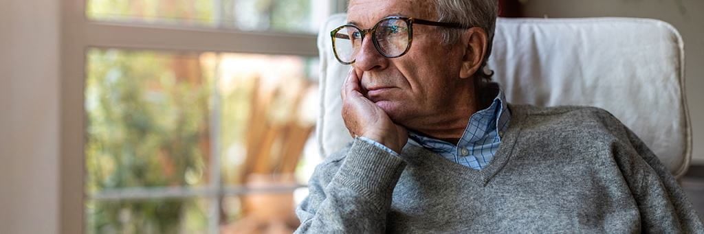 An anxious older man with glasses and a gray sweater looks out of a window.