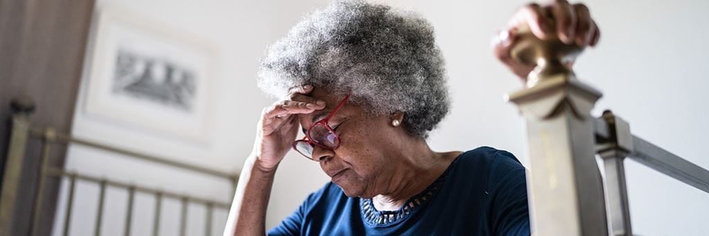 Older woman in blue shirt holding her head in pain while sitting on edge of bed.
