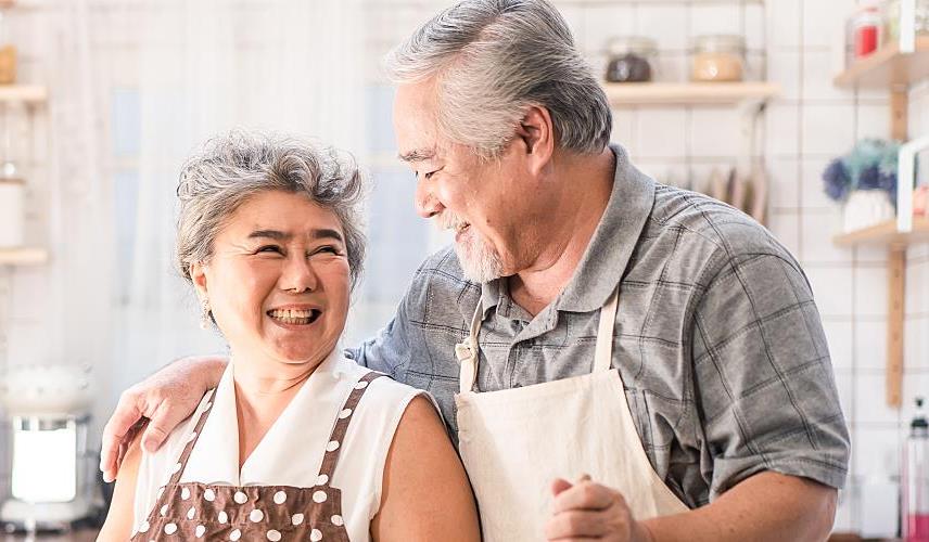 An older couple smiles at each other in their kitchen.