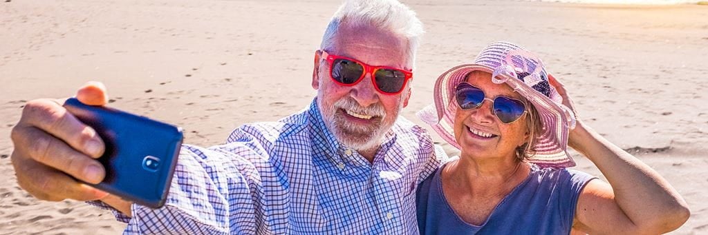 An older man in sunglasses takes a selfie on the beach with a woman in a sun hat.