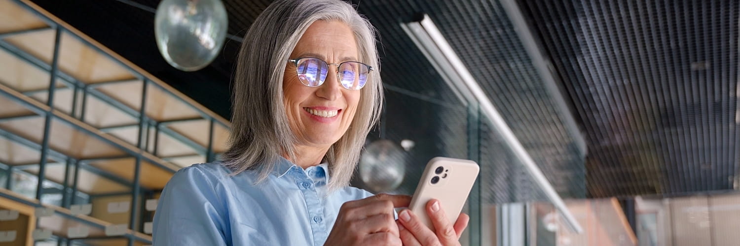 An older woman with gray hair smiles while using her phone.