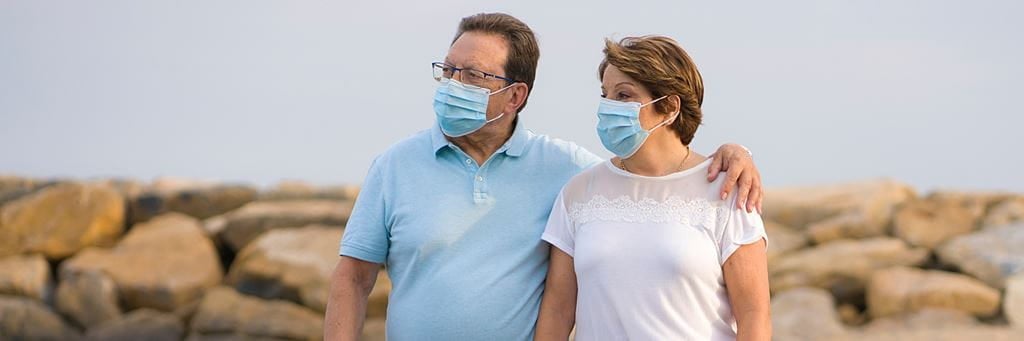 A retired couple walks on the beach while wearing face masks.