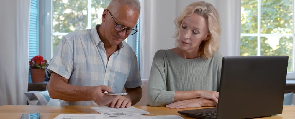 An older couple sits at a table with a laptop and financial documents.