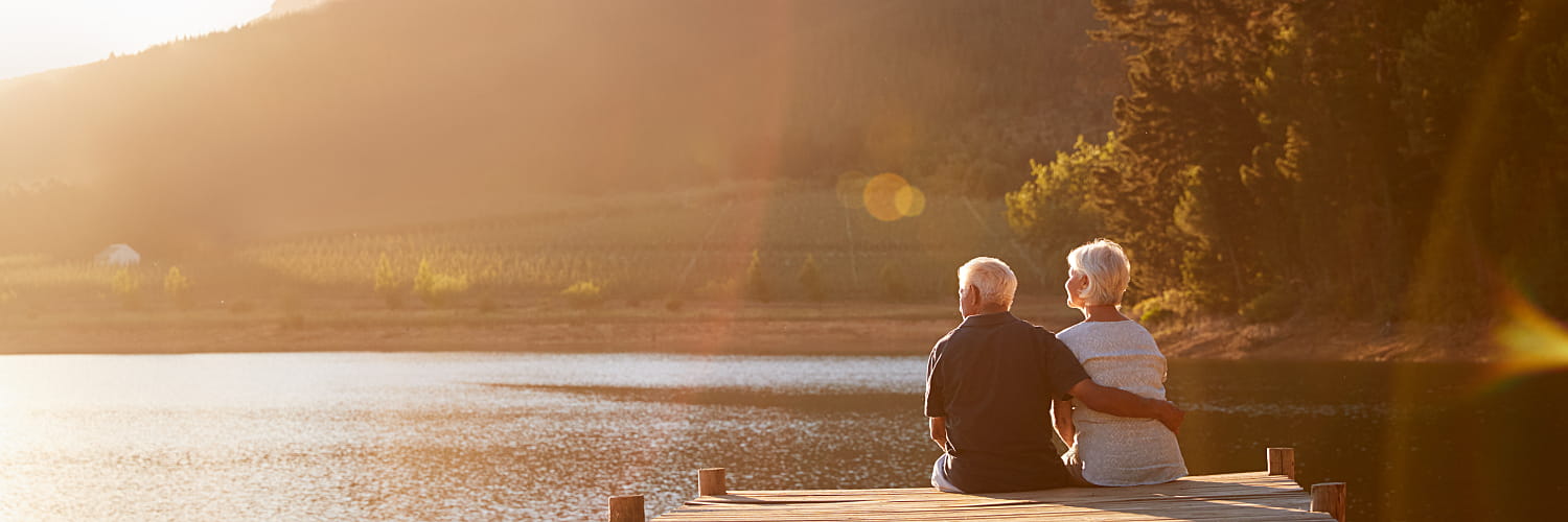 An older couple sits on a dock at the edge of a lake.