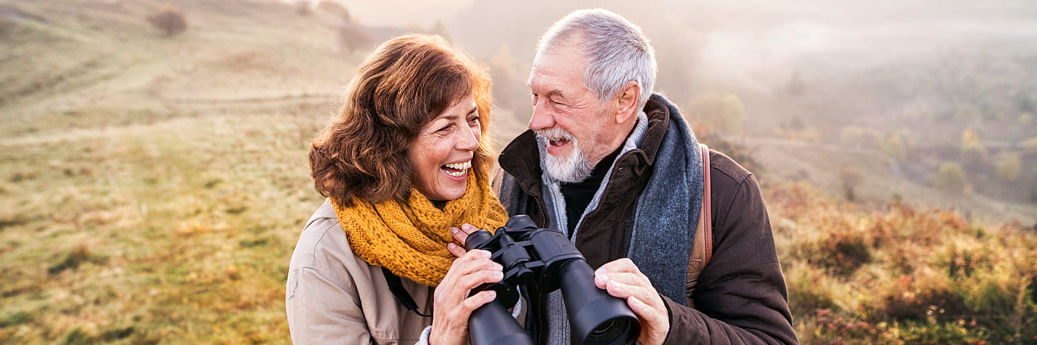 An older couple holding binoculars smiles at each other.