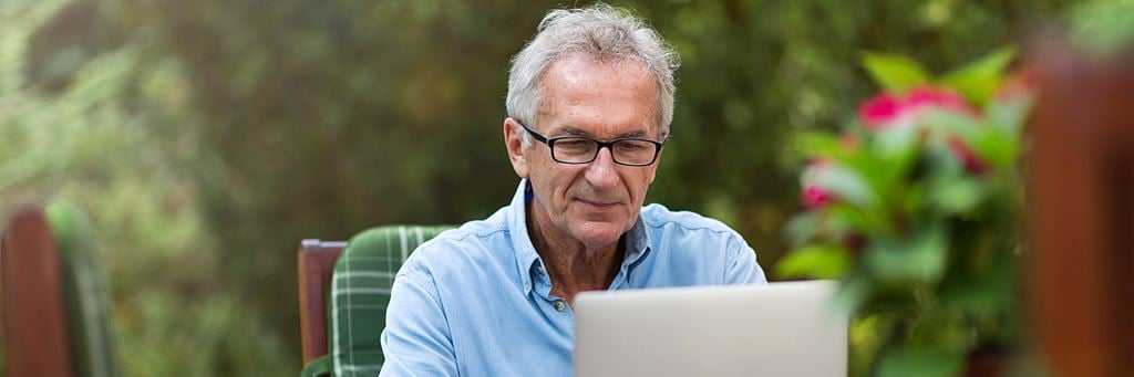 An older man works on a laptop while sitting in an outdoor patio.