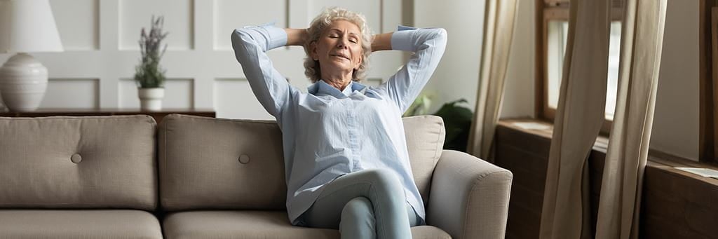 A senior woman relaxing on a couch with her hands behind her head.