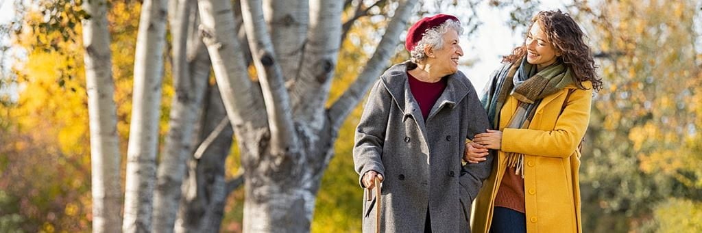 A senior woman and a younger woman wear warm clothing and walk in a park during fall.