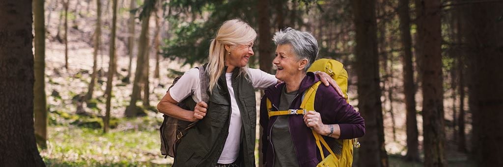 Retired women with white hair and backpacks walk together in the forest.