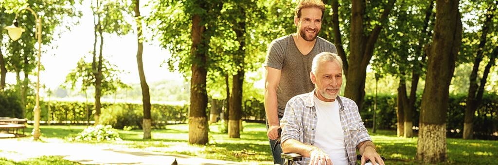 A middle-aged man walks in the park with his aging parent who is in a wheelchair.