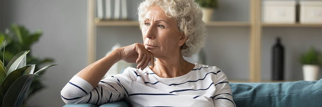 A thoughtful senior woman looks out the window while sitting on her living room couch.