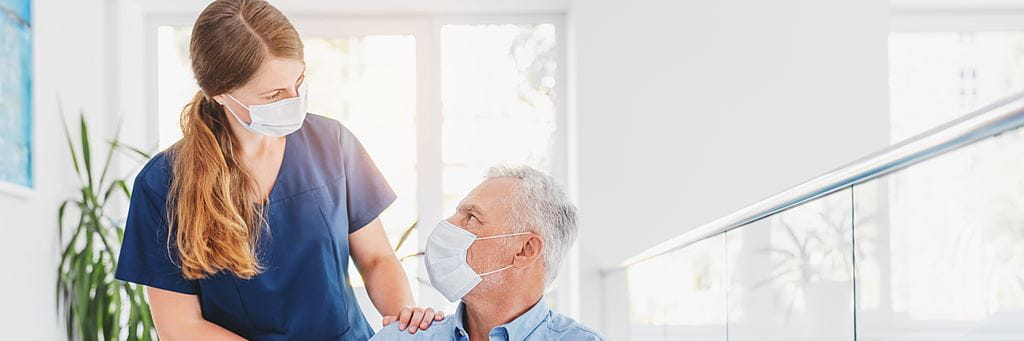 A medical professional wearing a mask talks to a senior patient wearing a mask.
